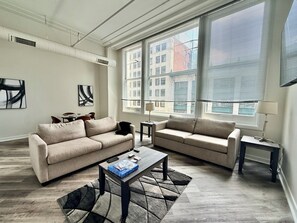 Ein modernes Wohnzimmer mit zwei Sofas, einem Couchtisch und einem Esstisch. Große Fenster bieten Blick auf die Stadtlandschaft.