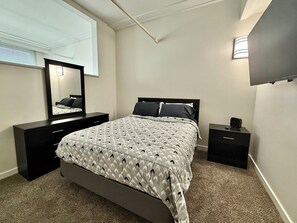 Ein Schlafzimmer mit Bett, Schminktisch, Nachttisch, Spiegel und an der Wand montiertem Fernseher.