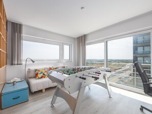 Ein modernes Zimmer mit einem Kicker, einer Couch mit bunten Kissen, einem blauen Schrank und großen Fenstern mit Blick auf die Stadt.