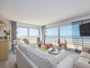 Een moderne woonkamer met een groot raam met uitzicht op een strand, een rond salontafeltje en een bank.