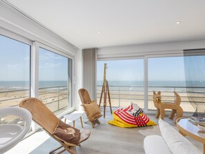 Ein geräumiges Zimmer mit großen Fenstern und Blick auf einen Strand, ausgestattet mit einer Couch, Stühlen und einer dekorativen Skulptur.