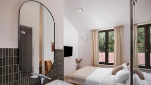 Quarto de hotel com um espelho grande, mesa de cabeceira, cama com almofadas, janela com cortinas e vista para o verde lá fora.