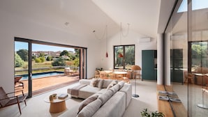 Uma sala de estar moderna com um sofá grande, área de refeições e vista para uma piscina e vegetação.