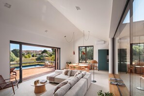 A modern living room with a large sofa, a dining area, and a view of a swimming pool and greenery. - Case Atrio Villas (Rovinj)