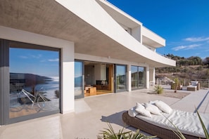 A modern house with large glass windows, a balcony with outdoor furniture, and a view of the sea.