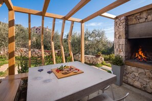 Un patio al aire libre con chimenea de piedra, una mesa con una bandeja de comida y copas de vino, y vistas del paisaje circundante.