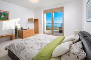 A bedroom with a bed, a bench, a dresser, a small table with a plant, and a painting on the wall.