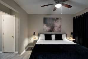 Un dormitorio con cama, ventilador de techo, un cuadro en la pared y dos mesitas de noche con lámparas.