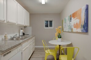 A kitchen with white cabinets, a granite countertop, a sink, and a window. There is a small table with yellow chairs and a vase of flowers. - The Palm Flats 11! Zoo! Museums! Medical Center! (Houston)