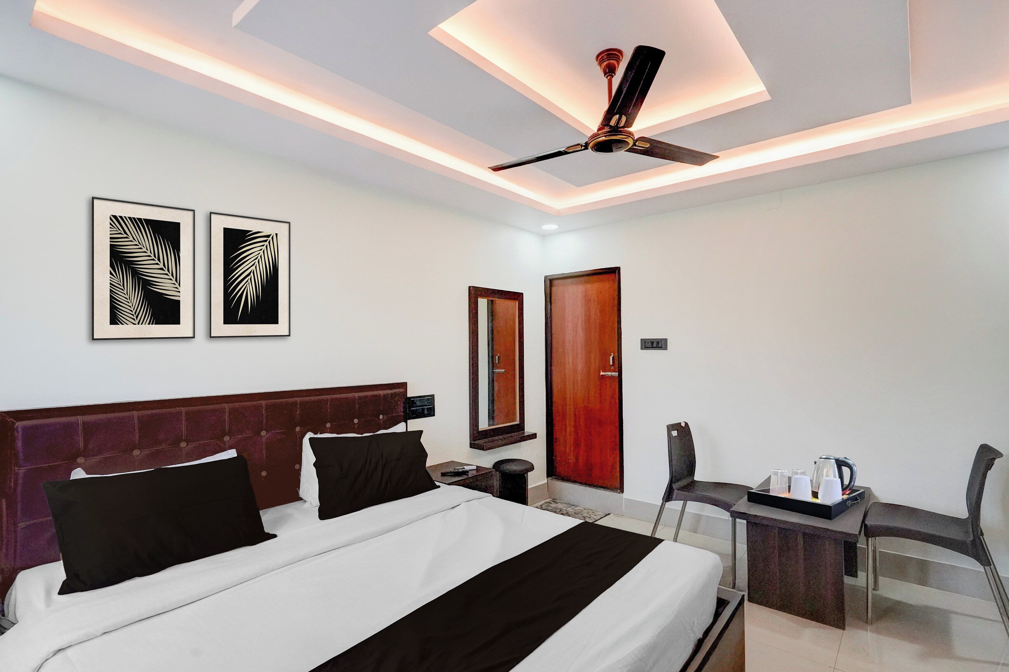 A hotel room with a bed, a desk with a chair, a ceiling fan, and two framed wall art pieces.