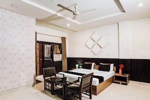 A hotel room with a bed, a desk, two chairs, a nightstand, a wall-mounted TV, and a ceiling fan.