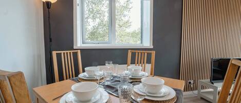 A dining table set with plates, bowls, glasses, and cutlery, with a window and blinds in the background.