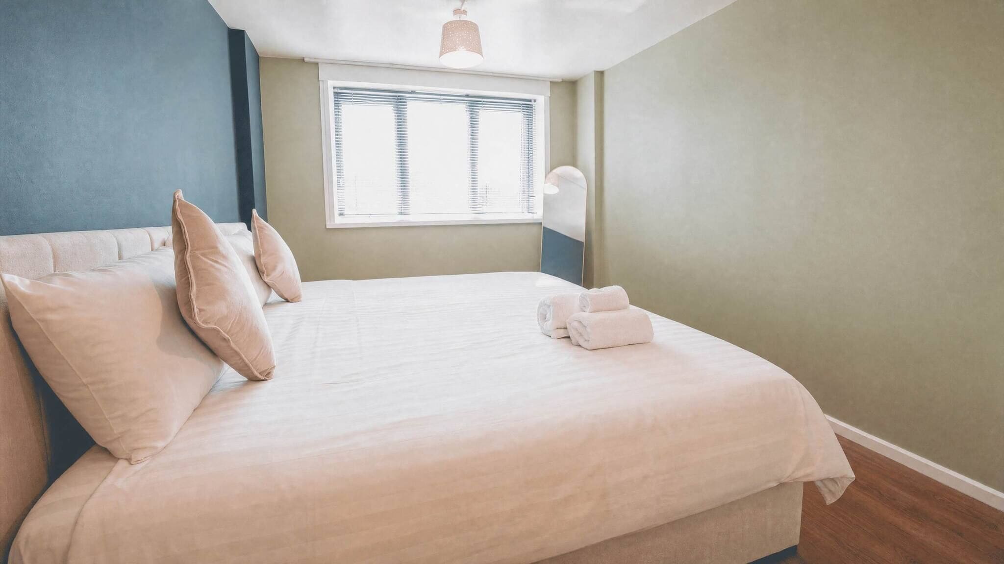 A neatly made bed with pillows and towels, a window with blinds, and a pendant light.