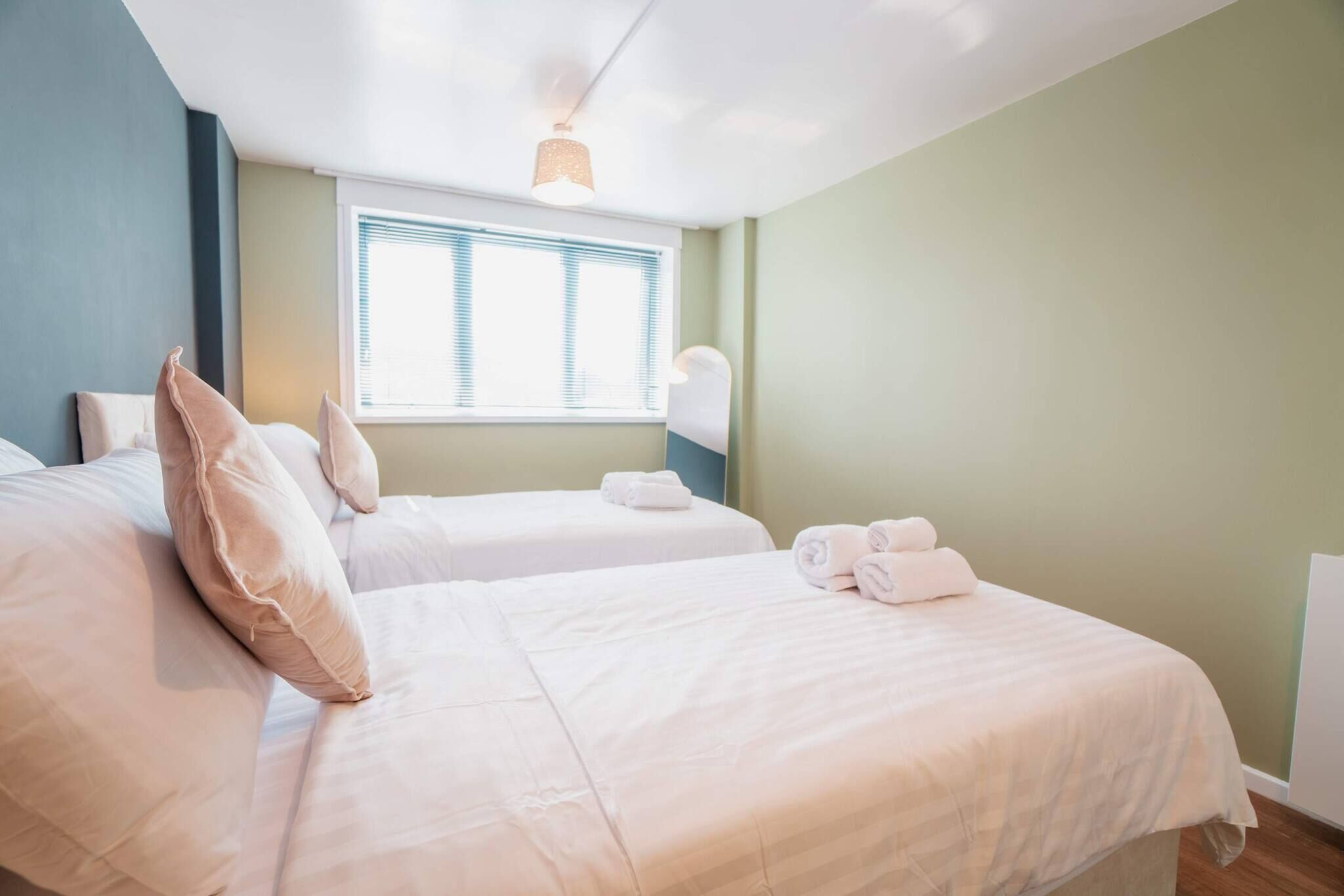 A hotel room with two beds, a window with blinds, a ceiling light, and a wall-mounted lamp.