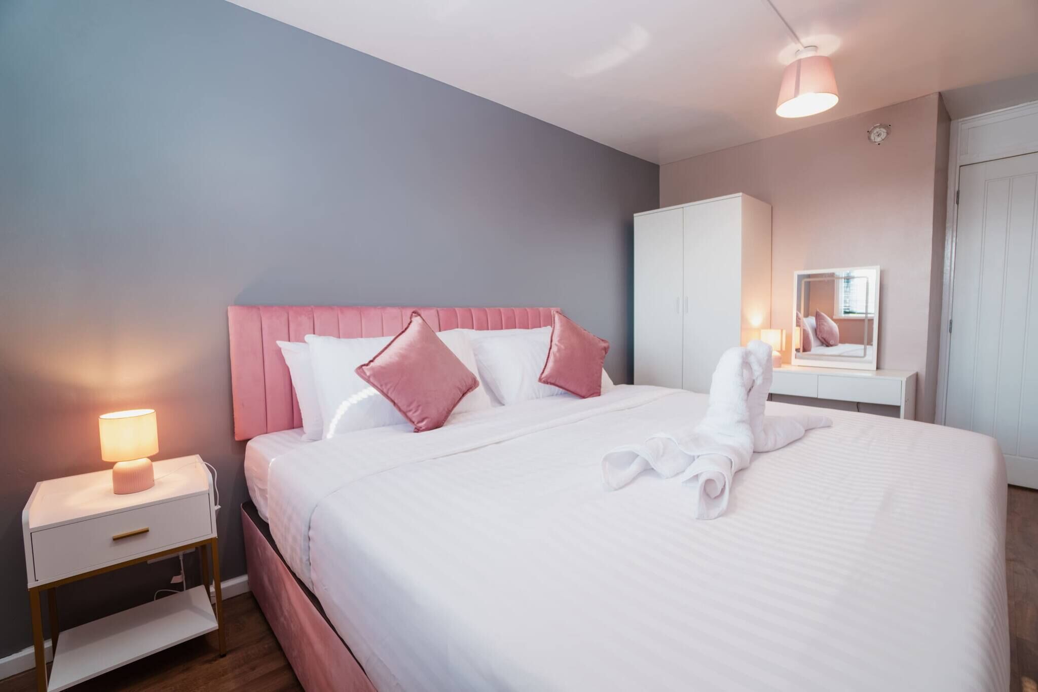 A neatly made bed with a pink headboard, a bedside lamp, and a mirror on the wall.