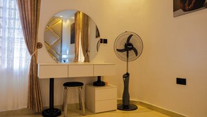 Una habitación con escritorio, espejo, ventilador de pie y un taburete.