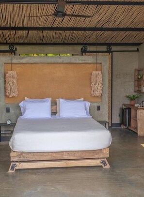 Una cama con estructura de madera y ropa de cama blanca, un ventilador de techo y un estante en la pared con una planta.