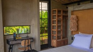 A bedroom with a bed, a desk with a chair, a wooden shelving unit, and a large window with a view of greenery.