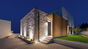 A modern two-story house with a stone facade and wooden paneling, featuring a well-lit entrance and a paved driveway. - Villa Rovni (Rasa)