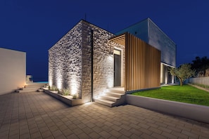 A modern two-story house with a stone facade and wooden paneling, featuring a well-lit entrance and a paved driveway. - Villa Rovni (Rasa)