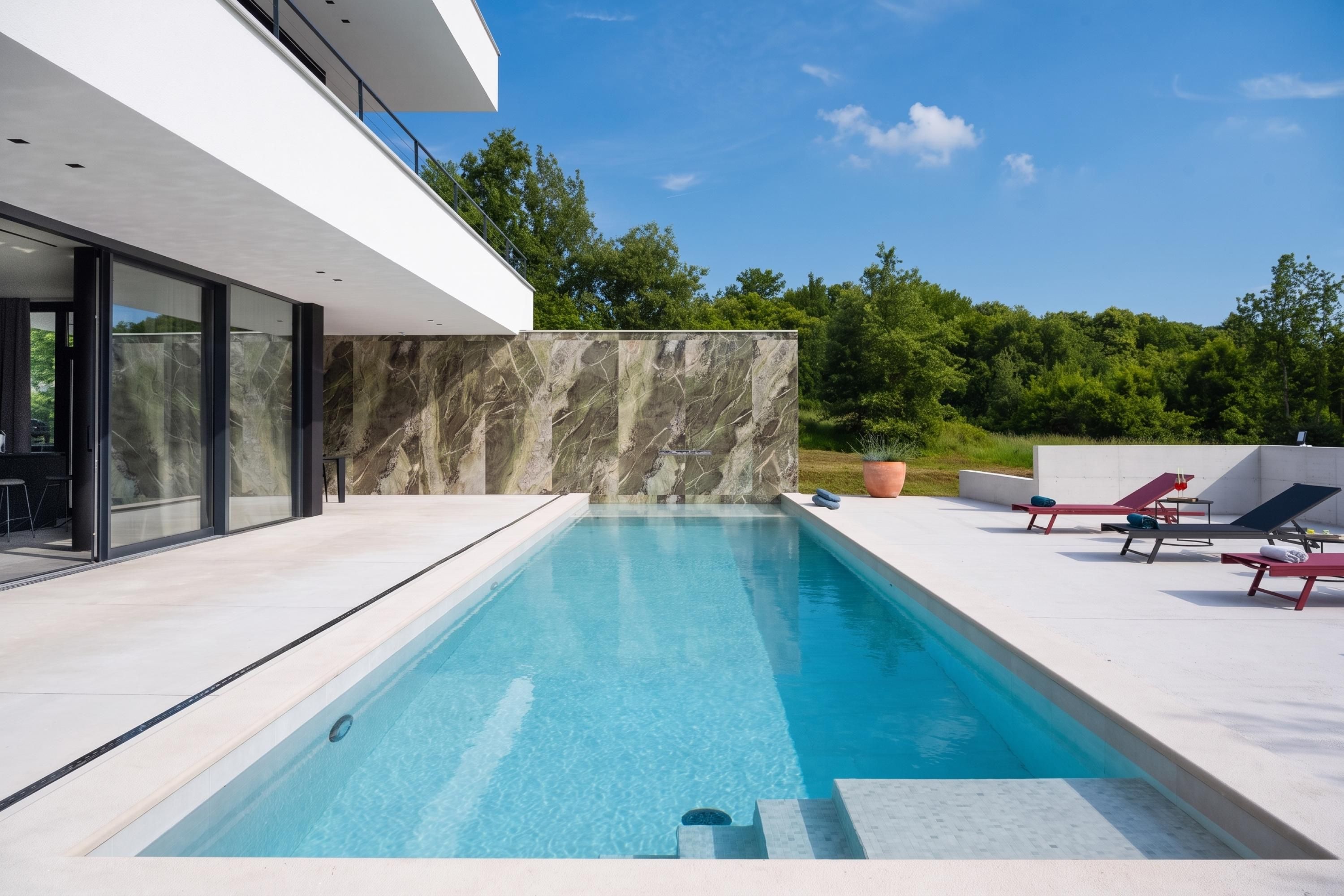 A modern outdoor swimming pool with a clear blue water surface, surrounded by a white deck and a stone wall.