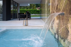 Ein moderner Außenpool mit Wasserfall, ein Essbereich mit Tischen und Stühlen sowie im Hintergrund eine Hecke und ein Gebäude.