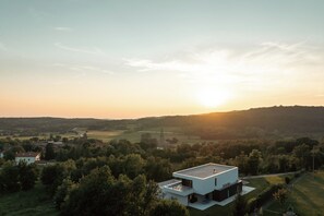 Ein modernes, zweistöckiges Haus mit flachem Dach, umgeben von einer üppigen, grünen Landschaft und blauem Himmel.