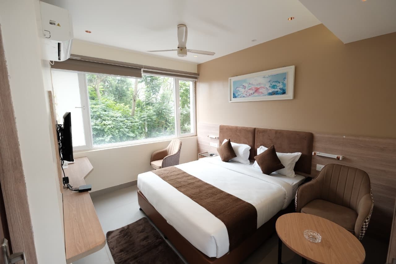 Habitación de hotel con cama, escritorio, silla, ventilador de techo y ventana con vistas a zonas verdes.