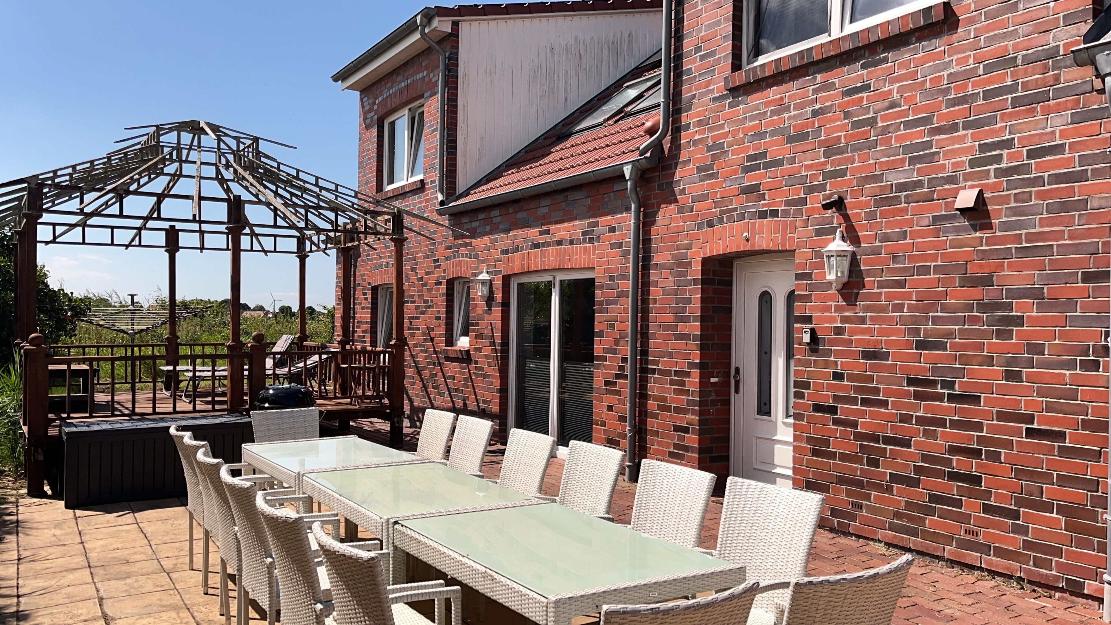 A patio with a glass-top table and wicker chairs, a brick building with a door and windows, and a metal pergola structure.