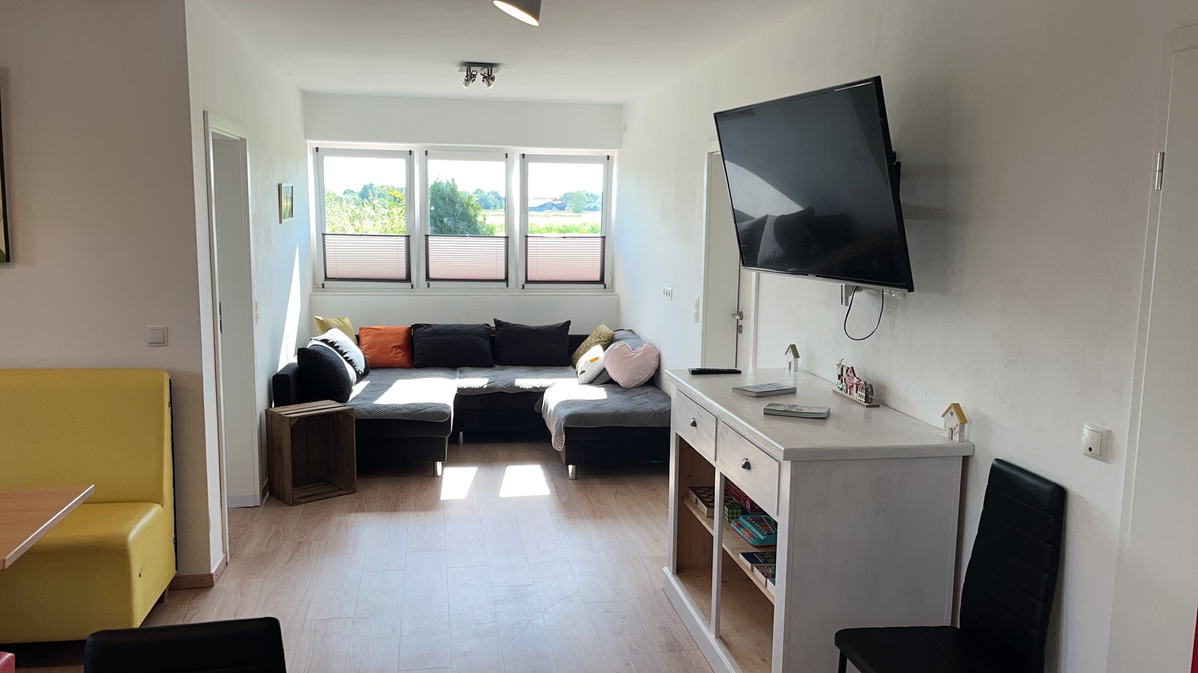 A modern living room with a sofa, a TV mounted on the wall, and a window with a view of greenery.