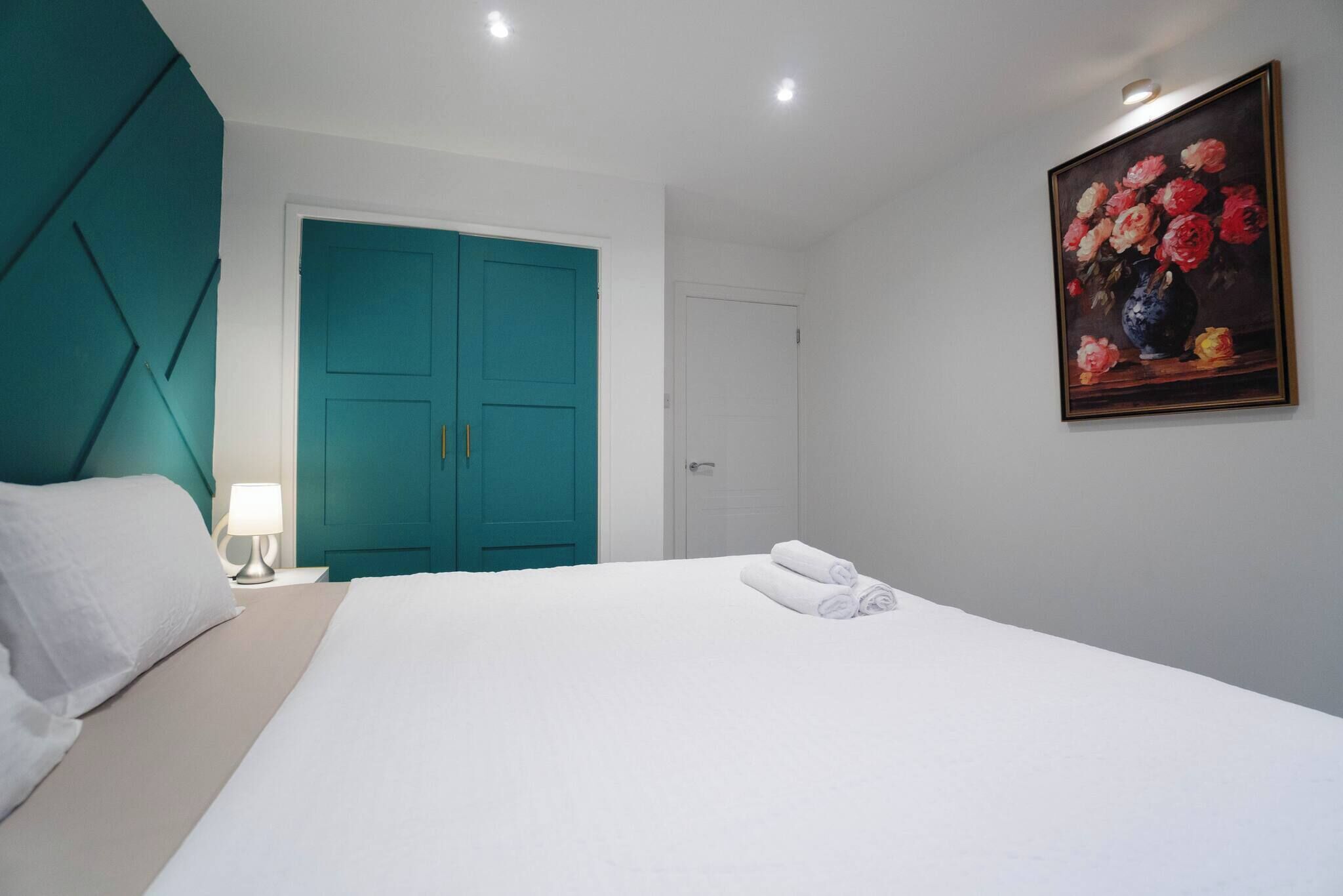 Habitación de hotel con una cama grande, un armario color aguamarina, un cuadro de flores en la pared y una mesita de noche con una lámpara.