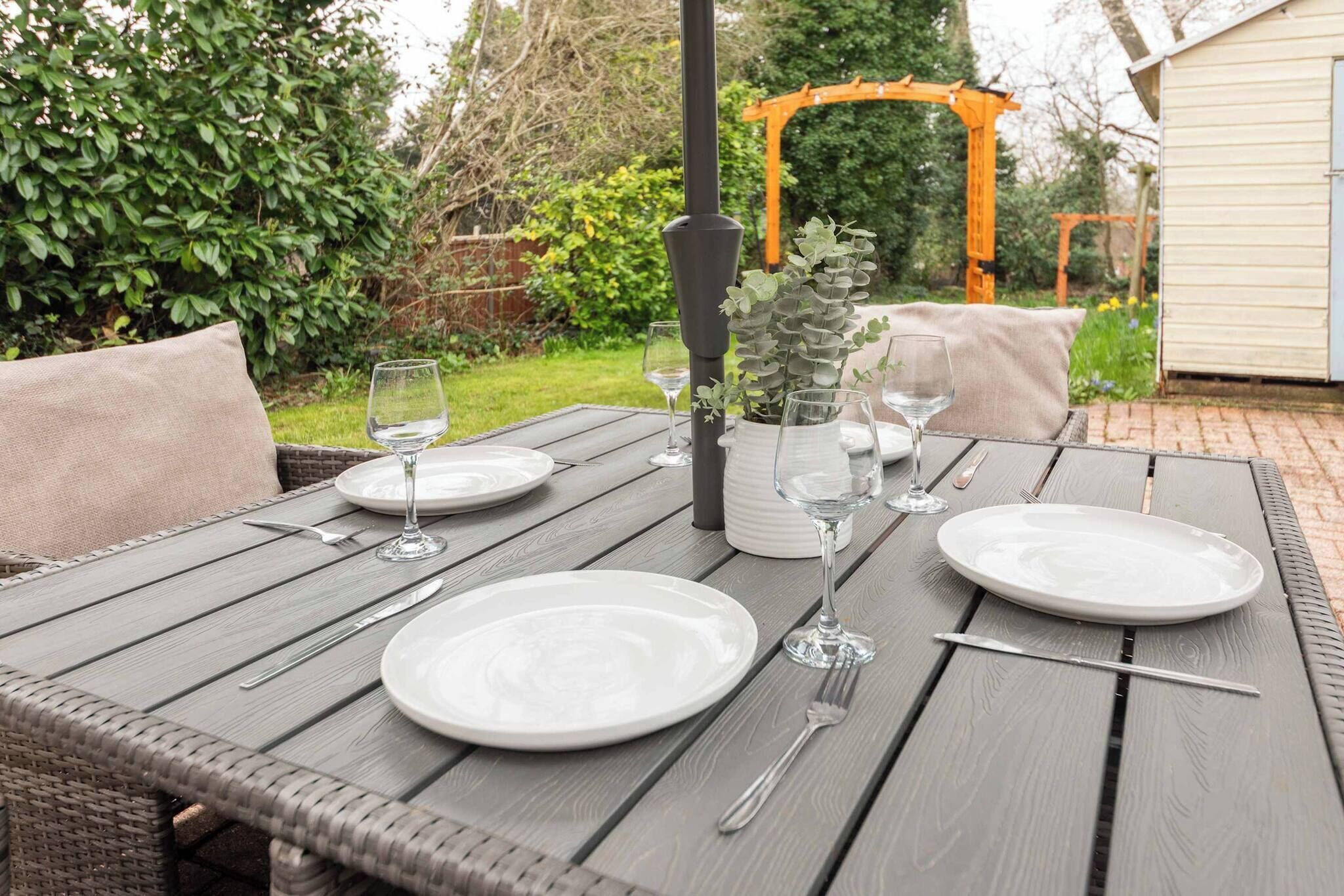 An outdoor dining table set with plates, glasses, and cutlery, surrounded by greenery and a wooden pergola.