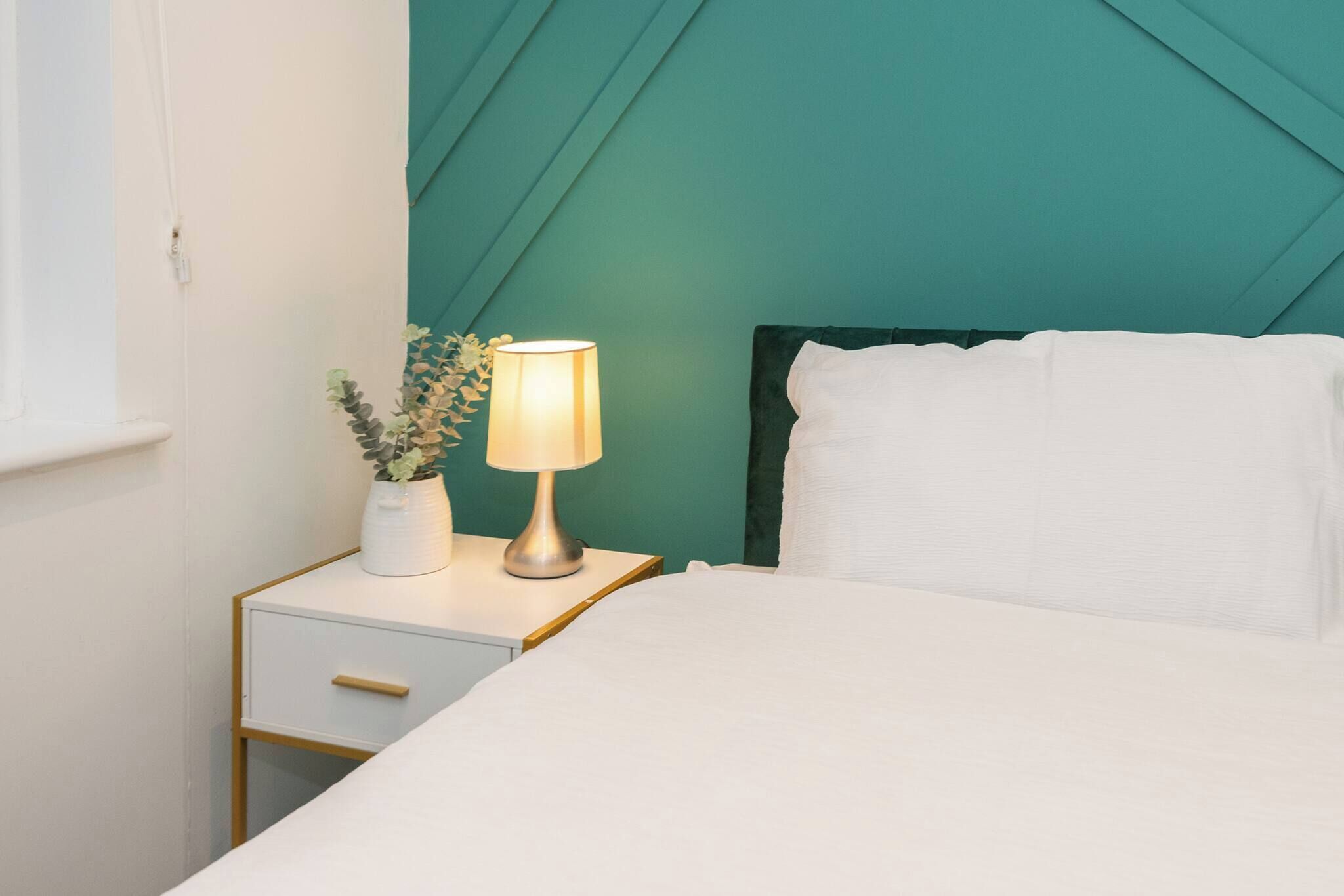 En pænt redt seng med hvide sengetøj, et natbord med en lampe og en lille potteplante.