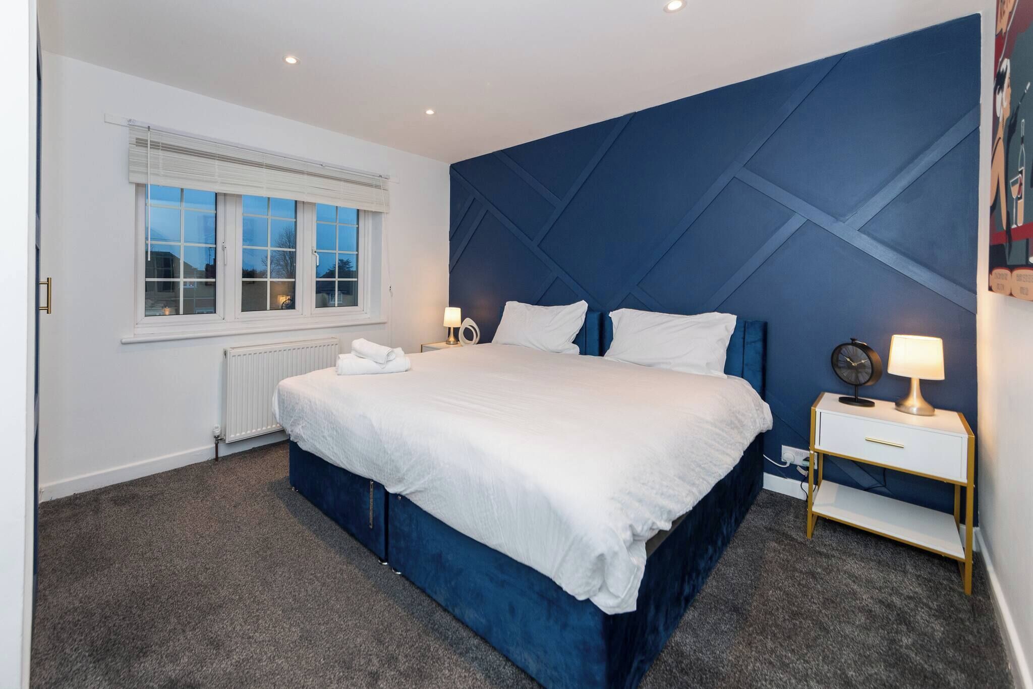 A modern bedroom with a large bed, a window with blinds, a radiator, a bedside table with a lamp, and a wall with a geometric pattern.
