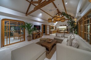Ein geräumiger Wohnbereich mit hohen Decken, Holzbalken, einer großen Sofagarnitur, einem Fernseher und einer Küche im Hintergrund.