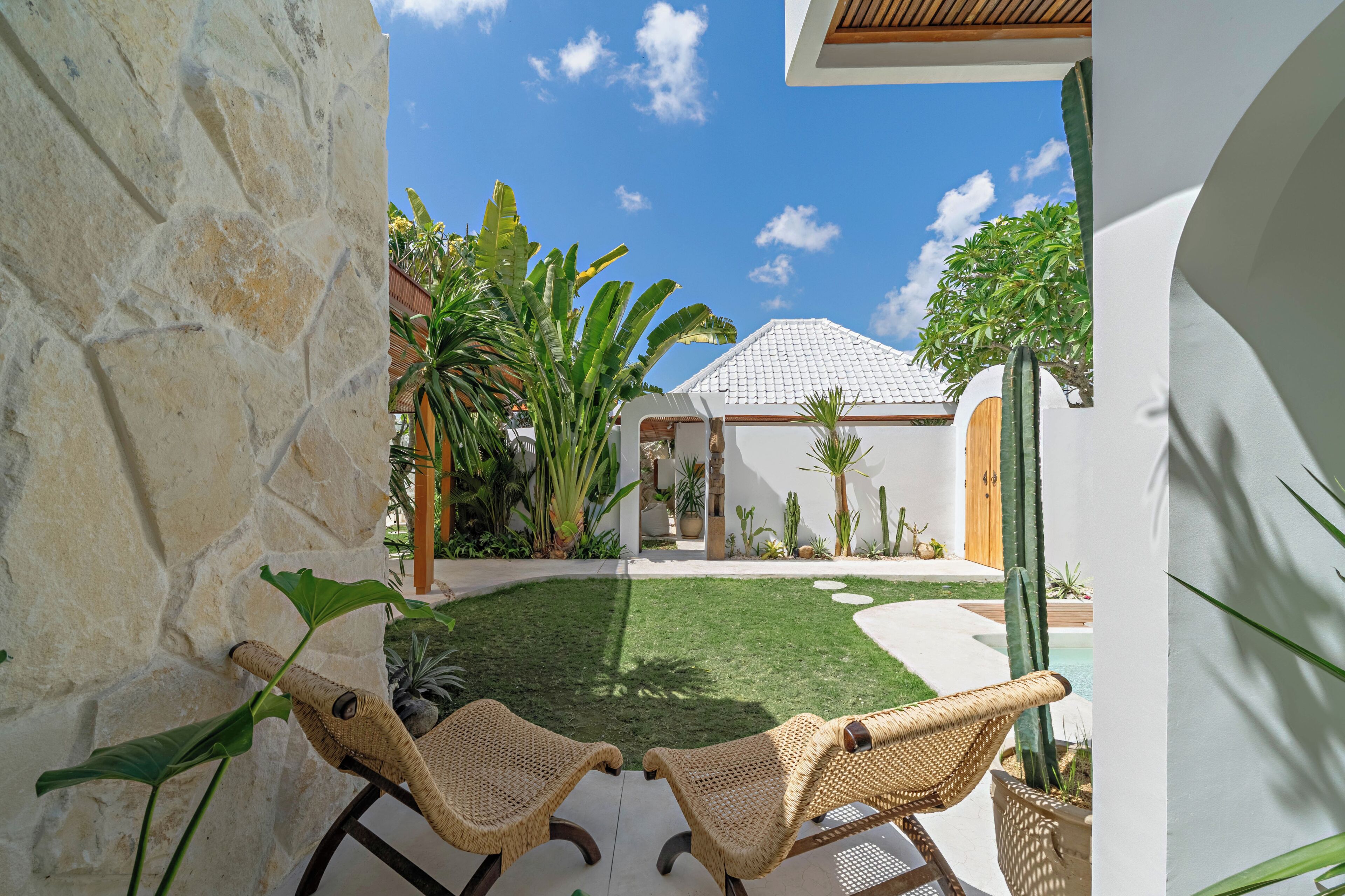 Ein Patio mit Steinmauer, zwei Korbstühlen, einer kleinen Rasenfläche und verschiedenen Pflanzen, darunter eine Bananenstaude und Kakteen.