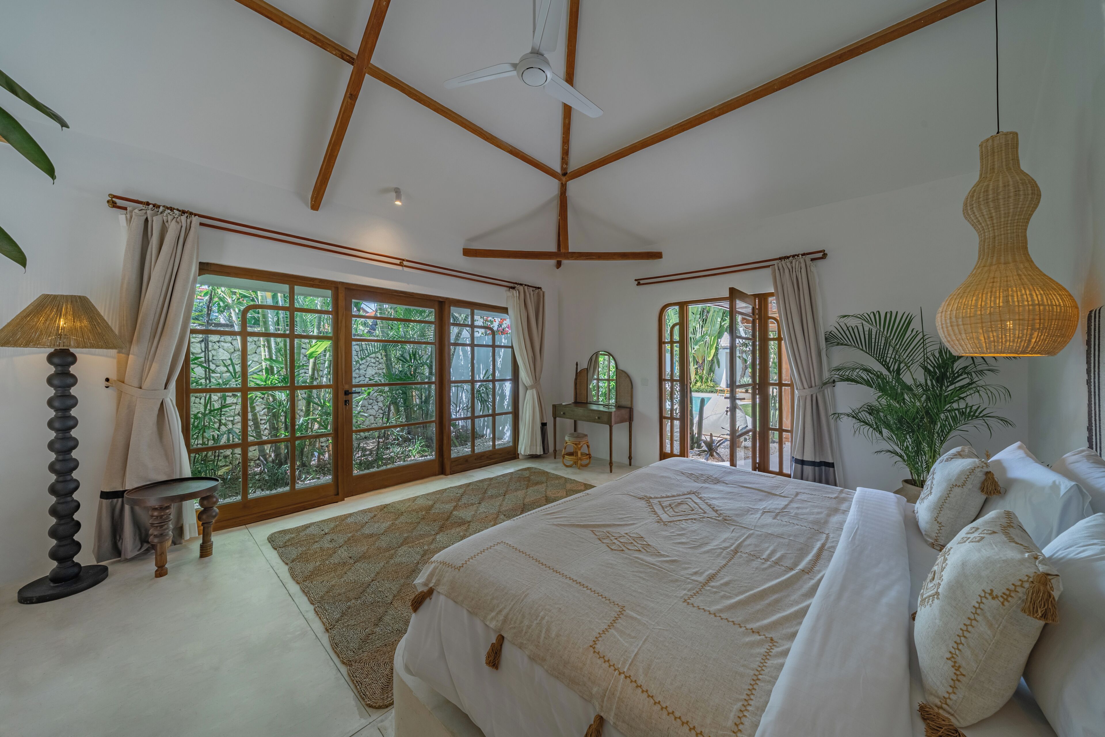 Une chambre spacieuse avec un grand lit, un ventilateur de plafond et une vue sur la verdure par les fenêtres.