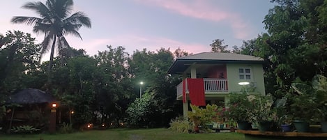 Uma casa com varanda, rodeada por vegetação exuberante e uma estrutura com telhado de palha.