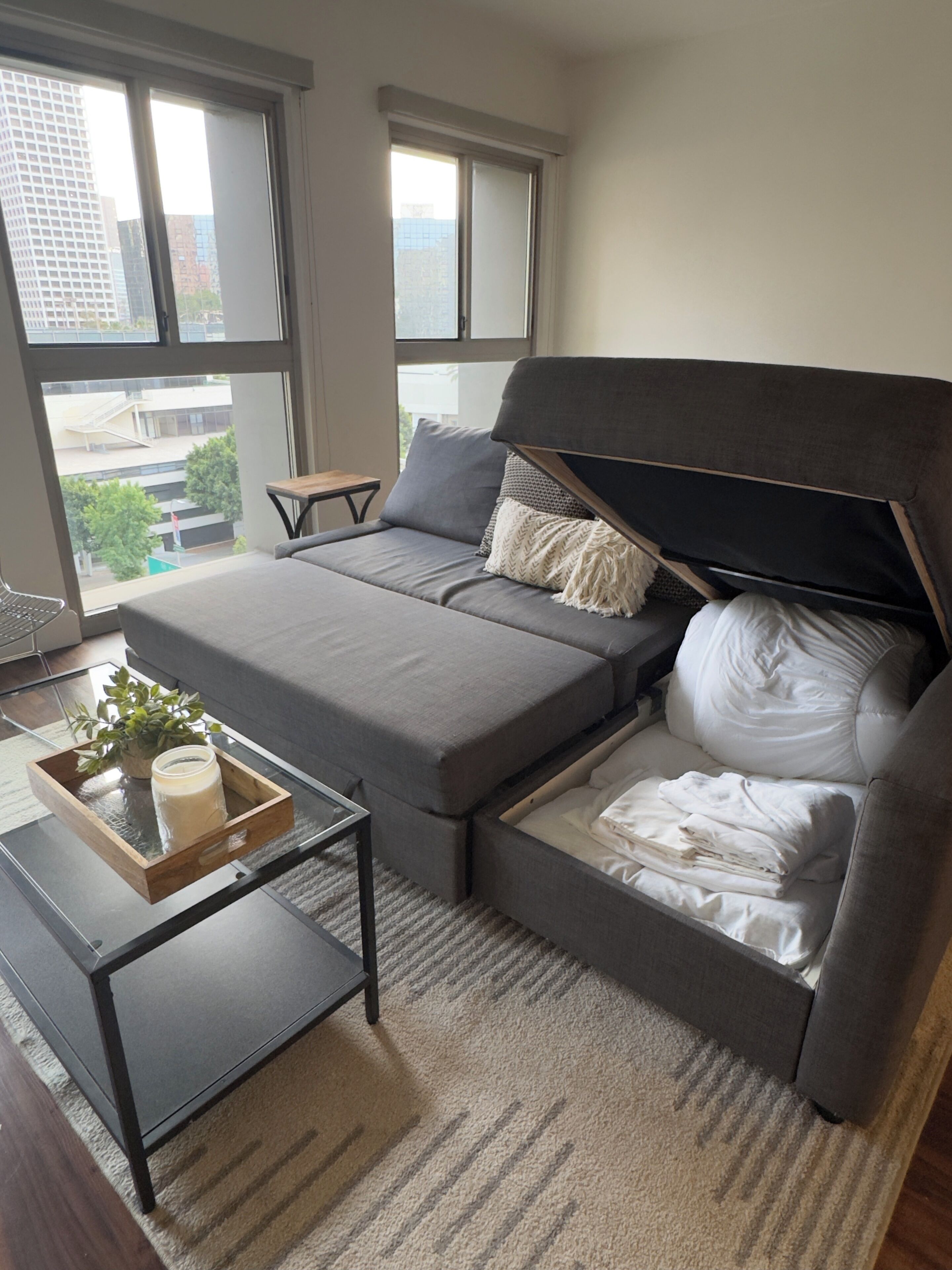 Ein modernes Schlafzimmer mit Schlafsofa, einem kleinen Tisch mit Pflanze und Blick auf die Stadt durch große Fenster.