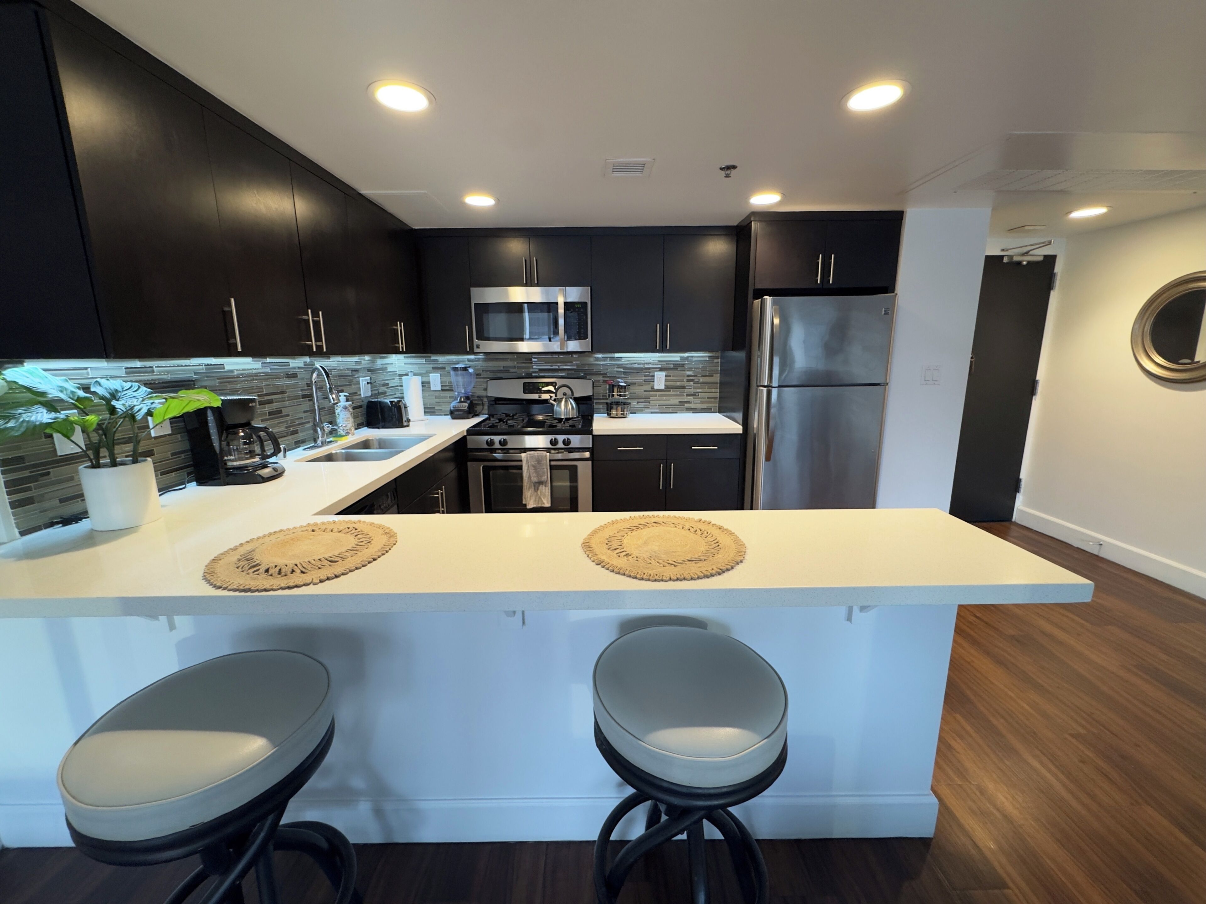A modern kitchen with dark cabinets, stainless steel appliances, a white island with bar stools, and a backsplash with a mosaic tile pattern.