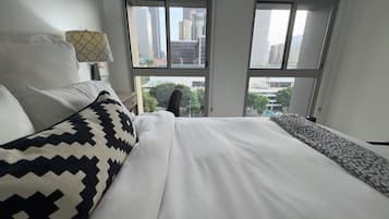 A hotel room with a large bed, two windows, a lamp, and a view of the cityscape.