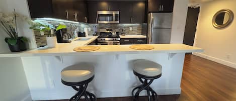 A modern kitchen with dark cabinetry, stainless steel appliances, a central island with stools, and a backsplash with a mosaic tile pattern.