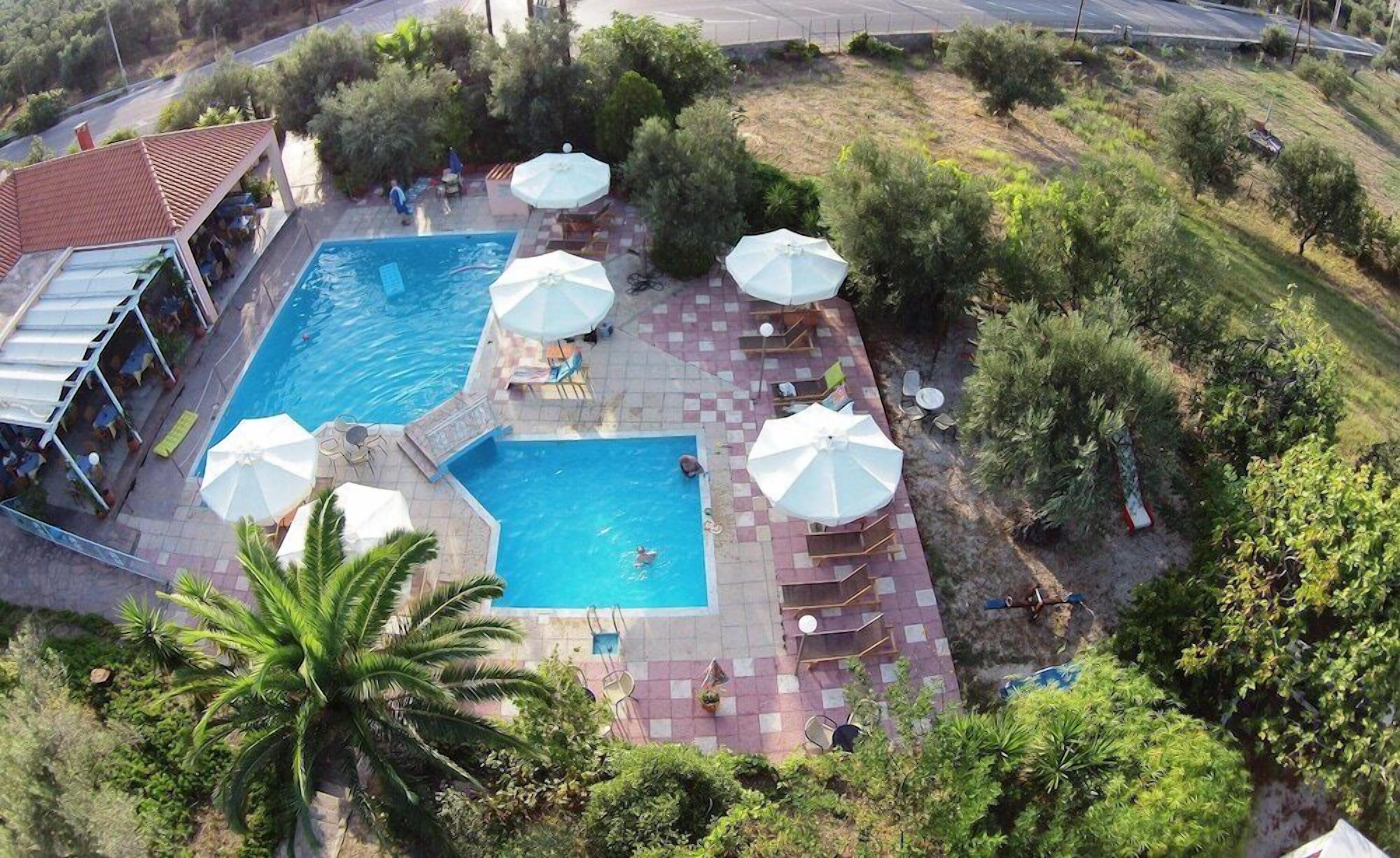 A swimming pool with lounge chairs and umbrellas, surrounded by greenery and a building with a red roof.