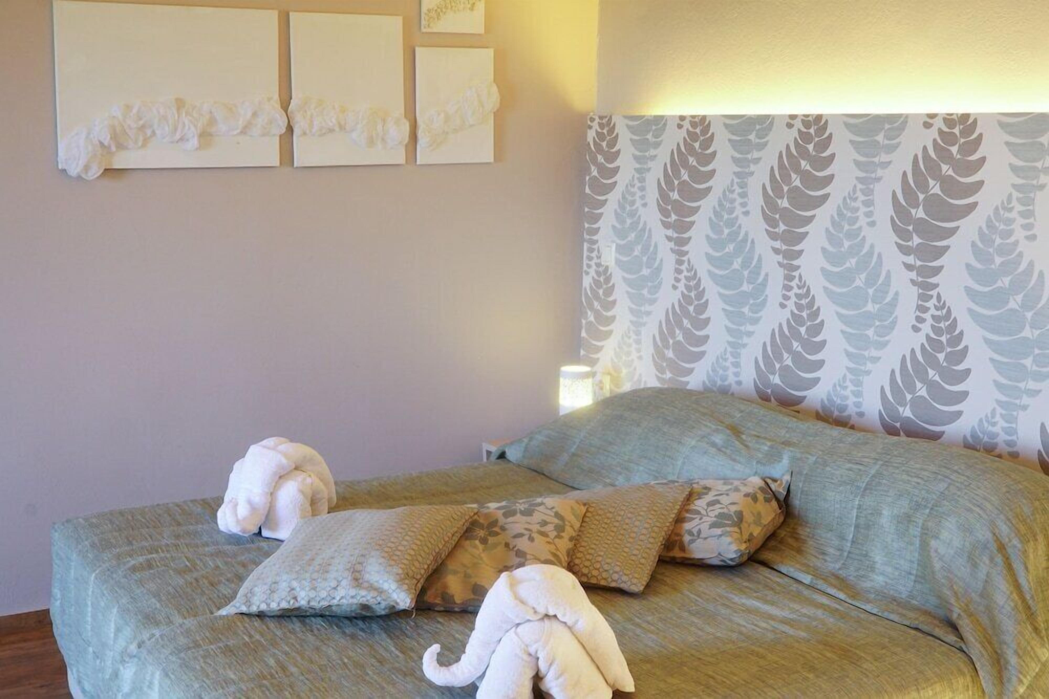 Una cama con dos almohadas y una toalla doblada, una pared con tres cuadros enmarcados, y un papel tapiz estampado.