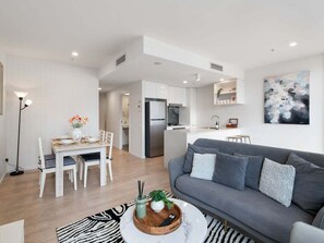 Ein modernes Wohnzimmer mit einem grauen Sofa, einem Esstisch mit Stühlen und einer Küche im Hintergrund.
