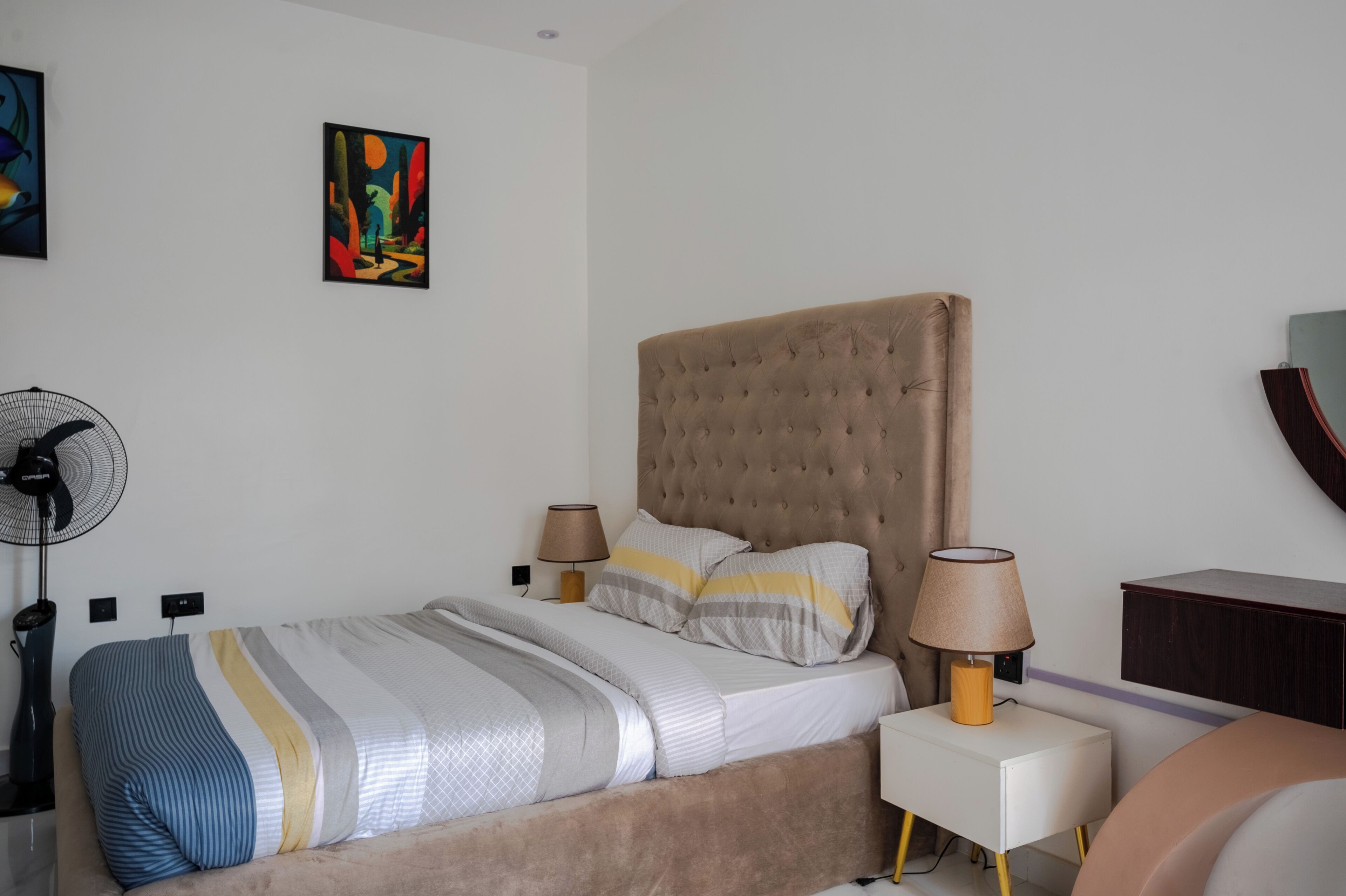 Una camera da letto ordinata con un letto, lampade sui comodini, una ventola e un quadro appeso al muro.