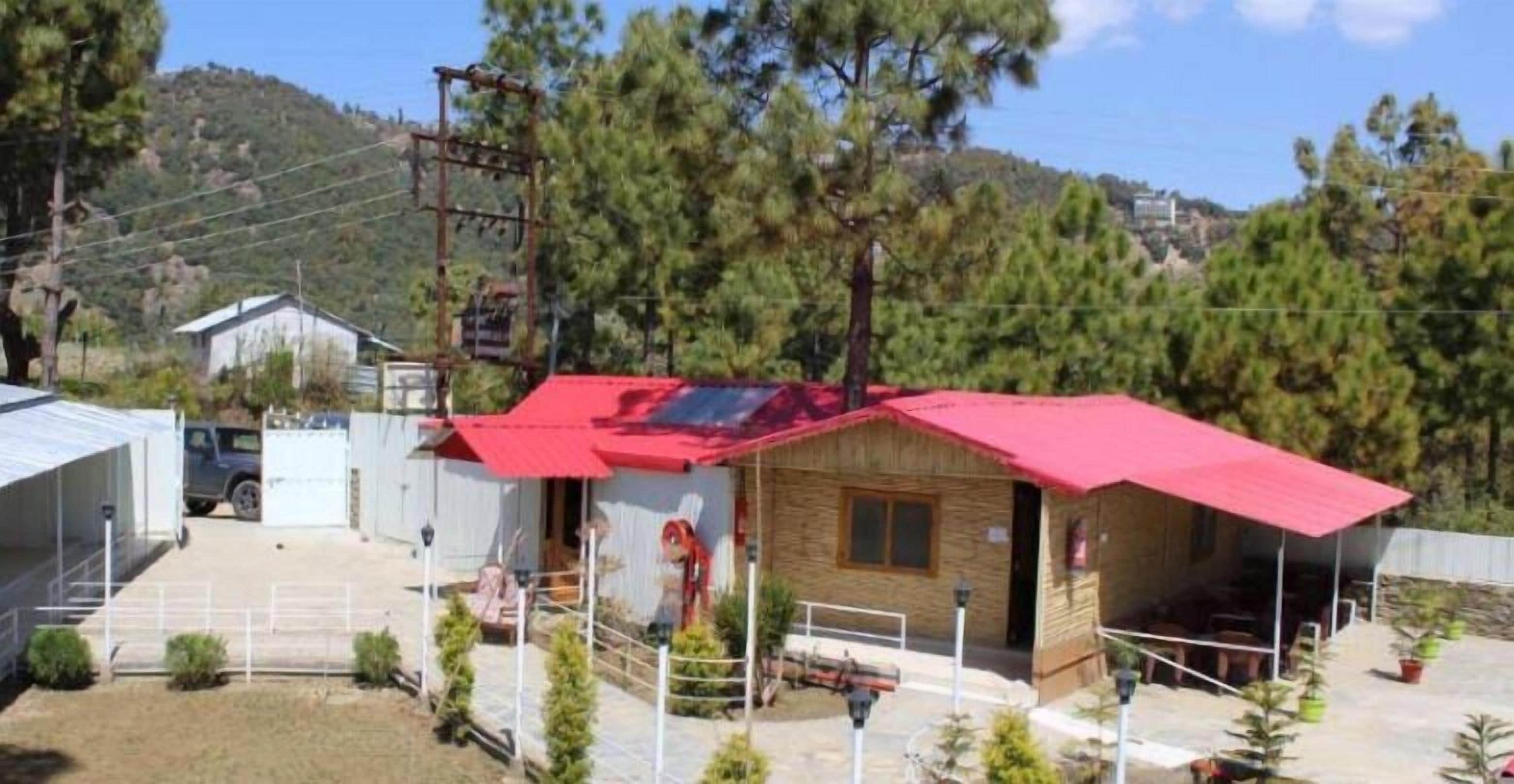 A small wooden cabin with a red roof, surrounded by a fenced area with potted plants, set against a backdrop of trees and hills.
