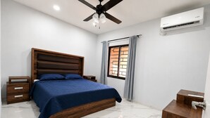 Un dormitorio con cama, mesitas de noche, ventilador de techo, equipo de aire acondicionado, ventana con cortinas y un armario de madera.