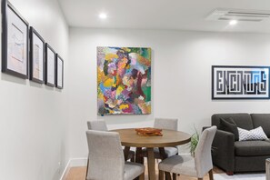A modern living room with a round wooden table, grey upholstered chairs, a grey sofa, and a colorful abstract painting on the wall. - NEW: Fun on 4th Encinitas (Aug 2026) rooftop fire pit (Encinitas)