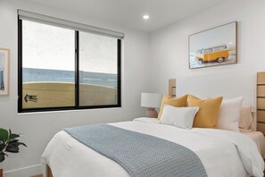 Ein Schlafzimmer mit großem Fenster, einem Bett mit vielen Kissen, einem Nachttisch mit Lampe und einem gerahmten Bild an der Wand.